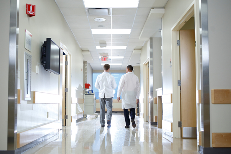 A low-angle view of two men in lab coats walking together down a sparkling clean, well-lit hallway. Light is bouncing off the mirrored finishes and streaming through the large windows backlighting the men.